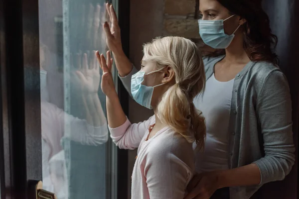 Foco seletivo de mãe e filha em máscaras médicas em pé perto da janela em casa — Fotografia de Stock