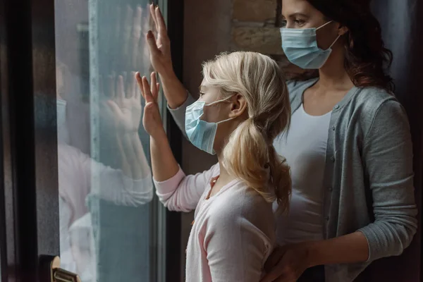 Selective focus of mother and daughter in medical masks standing near window at home — Stock Photo