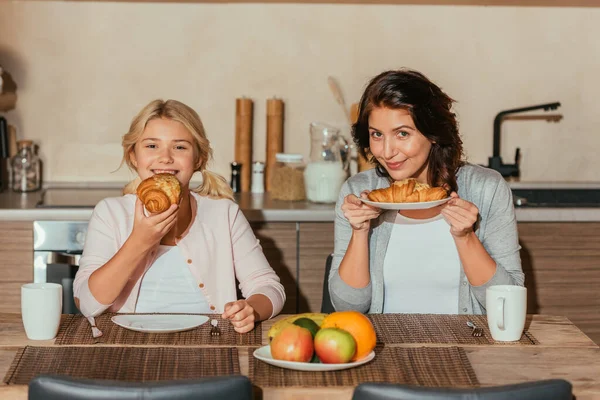 Selective focus of smiling child eating croissant near mother in kitchen — Stock Photo