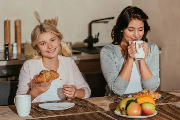 Lächelndes Kind blickt in die Kamera, während es in der Küche Croissant neben Mutter mit Tasse hält — Stockfoto