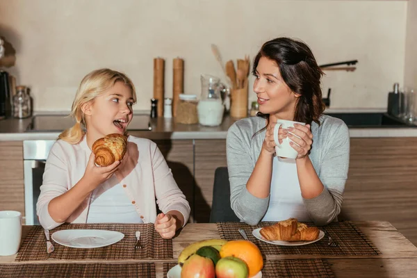 Selective focus of kid eating croissant and looking at smiling mother with cup in kitchen — Stock Photo