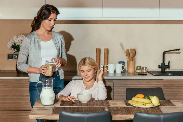 Criança sorridente sentada à mesa perto da mãe segurando frasco com cereais na cozinha — Fotografia de Stock