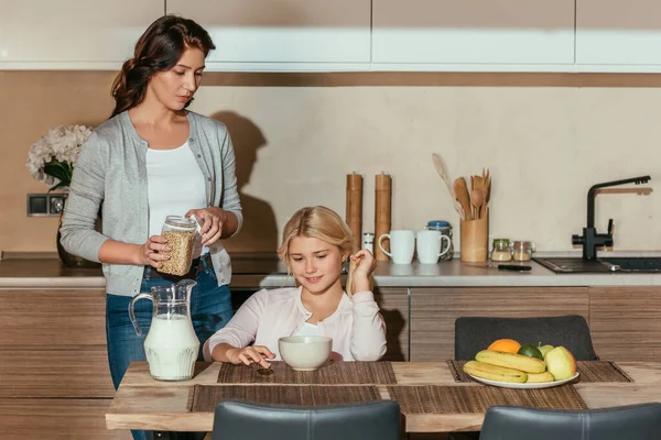 Niño sonriente sentado en la mesa cerca de la madre sosteniendo frasco con cereales en la cocina - foto de stock