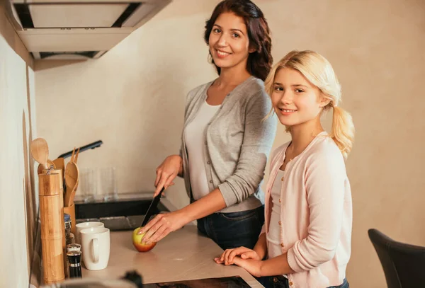 Seitenansicht von lächelnder Mutter und Tochter, die in die Kamera schauen, während sie Apfel auf Küchenarbeitsplatte schneiden — Stockfoto