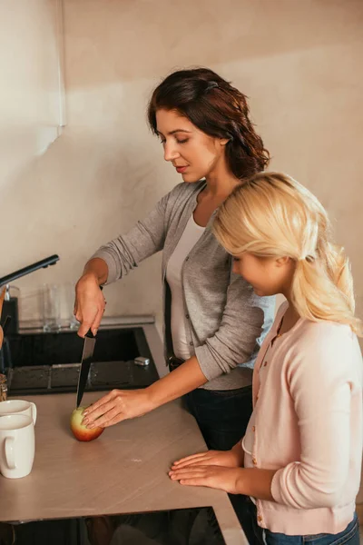Side view of daughter looking at mother cutting apple on kitchen worktop — Stock Photo