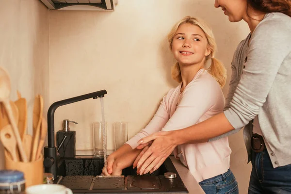 Foco seletivo de criança sorridente lavar as mãos perto da mãe na cozinha — Fotografia de Stock