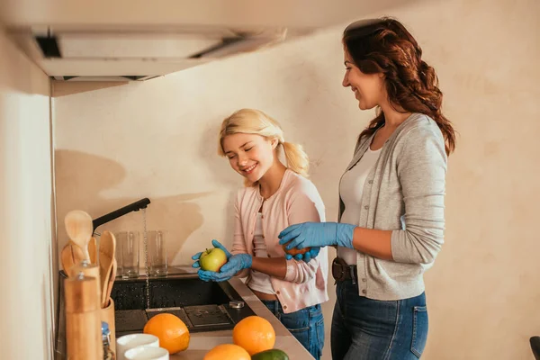 Selektiver Fokus lächelnder Mutter und Kind in Latex-Handschuhen beim Obstwaschen in der Küche — Stockfoto