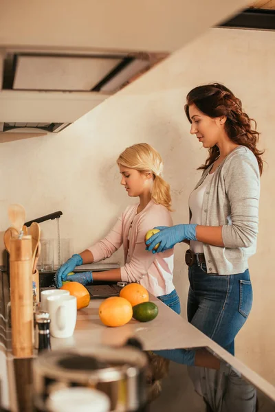 Vista laterale di madre e figlia in guanti di lattice lavare frutta in cucina — Foto stock