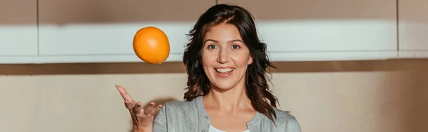 Панорамный урожай улыбающейся женщины, бросающей апельсин на кухню — стоковое фото