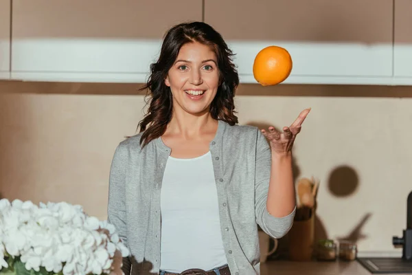 Selective focus of smiling woman throwing orange near flowers in kitchen — Stock Photo
