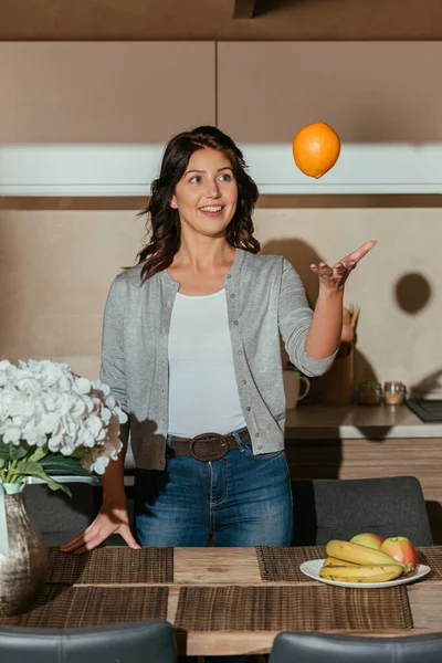 Selective focus of beautiful smiling woman throwing orange near flowers and fruits on kitchen table — Stock Photo