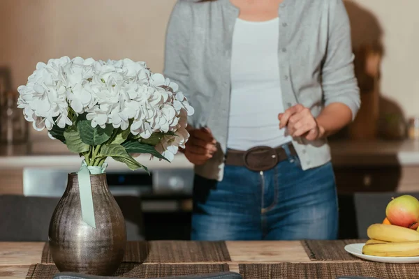 Foco seletivo de flores em vaso e frutas na mesa perto de mulher na cozinha — Fotografia de Stock