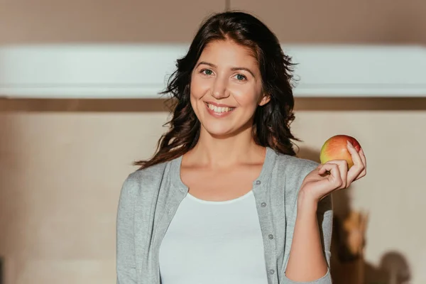 Schöne lächelnde Frau blickt in die Kamera, während sie frische Äpfel in der Küche hält — Stockfoto