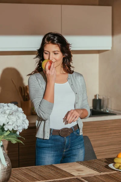 Selective focus of beautiful woman smelling apple and looking at flowers on kitchen table — Stock Photo