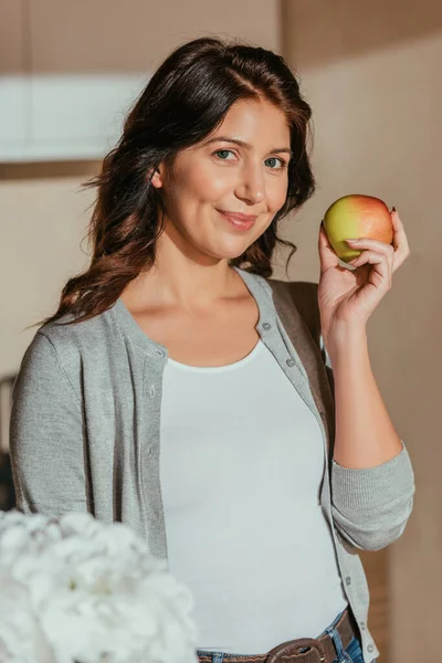 Selective focus of beautiful woman smiling at camera while holding organic apple in kitchen — Stock Photo