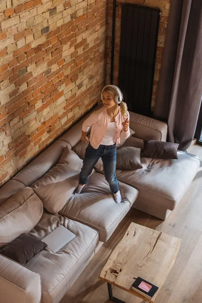 Vue grand angle de l'enfant gai dans les écouteurs debout sur le canapé près de l'ordinateur portable à la maison — Photo de stock