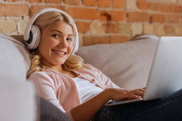 Selective focus of smiling kid in headphones using laptop on sofa — Stock Photo