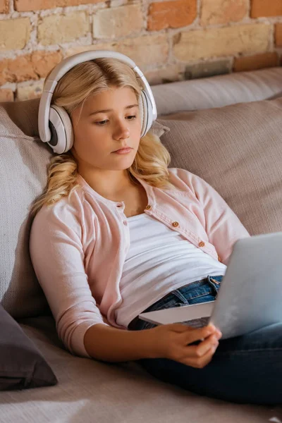 Concentration sélective de l'enfant dans les écouteurs regardant l'ordinateur portable sur le canapé à la maison — Photo de stock