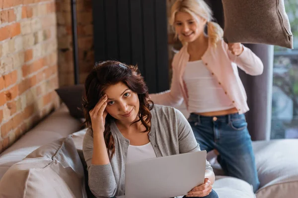 Selektiver Fokus einer Frau mit Laptop, die in die Kamera lächelt, in der Nähe eines Kindes, das Kissenschlacht auf der Couch spielt — Stockfoto