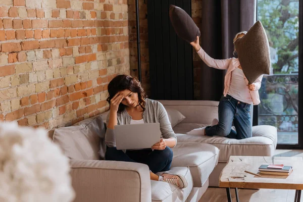 Selective focus of woman working on laptop near daughter playing pillow fight on couch — Stock Photo