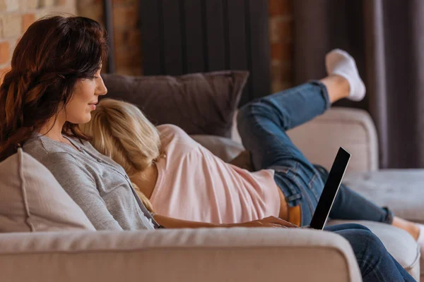 Selective focus of woman using laptop near daughter on sofa in living room — Stock Photo