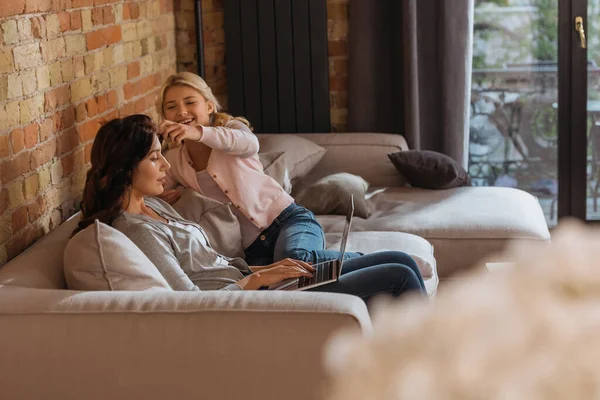 Выборочный фокус улыбающейся дочери трогательной матери, работающей на диване дома на ноутбуке — стоковое фото