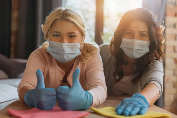 Selektiver Fokus des Kindes in Latex-Handschuhen zeigt Daumen hoch neben Mutter in medizinischer Maske, die Lappen im Wohnzimmer hält — Stockfoto