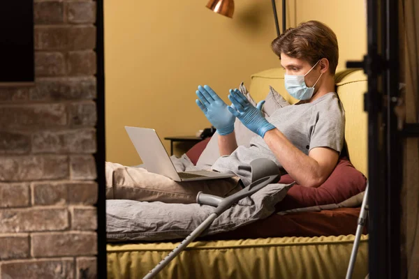 Foco seletivo do homem com deficiência em máscara médica e luvas de látex com bate-papo por vídeo no laptop no quarto — Fotografia de Stock