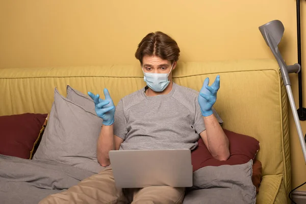 Homem com deficiência em máscara médica com videochamada no laptop perto da muleta no quarto — Fotografia de Stock