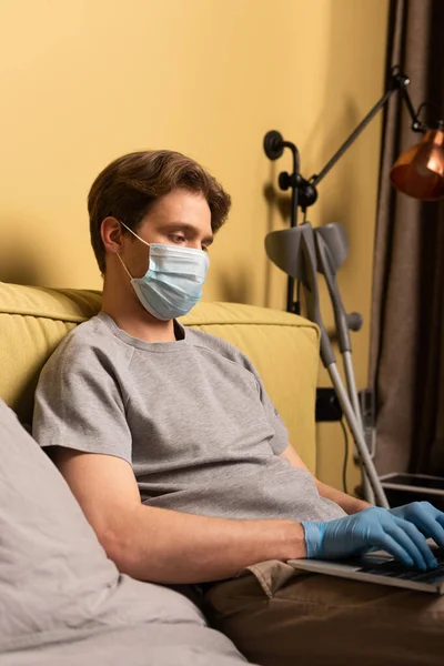 Man in medical mask and latex gloves using laptop near crutches in bedroom — Stock Photo