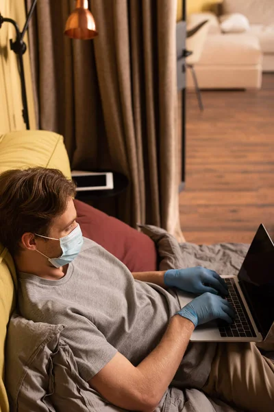 Uomo in maschera medica e guanti in lattice utilizzando il computer portatile sul letto — Foto stock