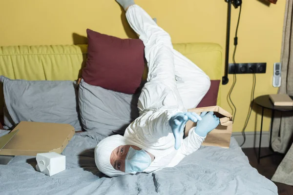Mann in Schutzanzug, Schutzmaske und Latexhandschuhen zieht Hand und hält Paket auf dem Bett — Stockfoto