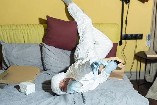 Man in hazmat suit, medical mask and latex gloves pulling hand and holding package on bed — Stock Photo