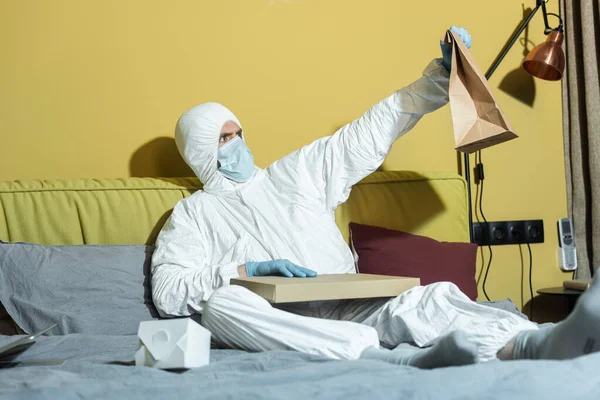 Foco seletivo do homem em terno hazmat e máscara médica segurando pacote perto da caixa de pizza na cama — Fotografia de Stock