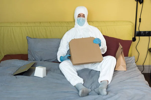 Uomo in tuta hazmat, guanti in lattice e maschera medica che tiene la scatola della pizza vicino ai pacchetti sul letto — Foto stock