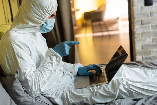 Side view of man in hazmat suit, medical mask and latex gloves pointing with finger at laptop on bed — Stock Photo