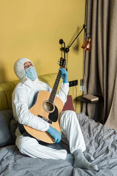 Homme en combinaison Hazmat, masque médical et gants en latex tenant une guitare acoustique au lit — Photo de stock