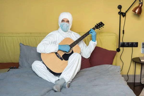 KYIV, UCRANIA - 24 DE ABRIL DE 2020: Hombre con traje de materiales peligrosos y máscara médica sosteniendo la guitarra acústica y mirando a la cámara en el sofá - foto de stock