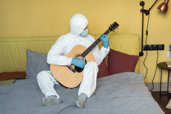 KYIV, UCRANIA - 24 DE ABRIL DE 2020: Hombre con traje de hazmat y máscara médica tocando la guitarra acústica cerca de la computadora portátil y joystick en la cama - foto de stock