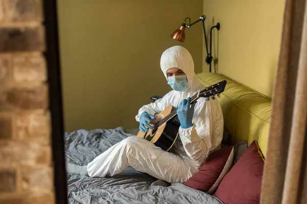 Selective focus of man in hazmat suit and latex gloves playing acoustic guitar in bedroom — Stock Photo