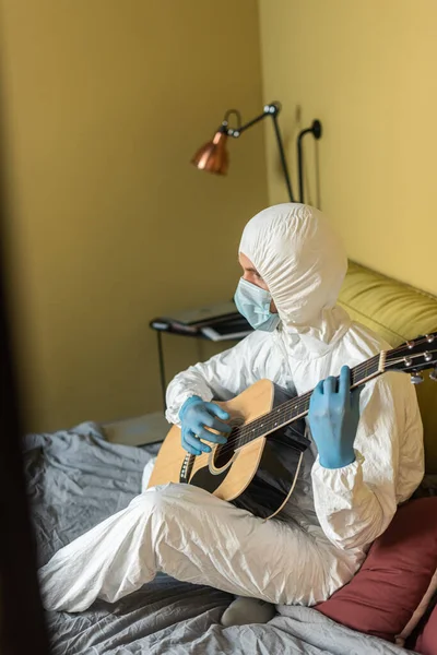 Vue latérale de l'homme en costume hazmat jouant de la guitare acoustique sur le lit à la maison — Photo de stock