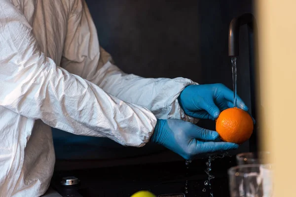 Cropped view of man in latex gloves and hazmat suit washing orange in kitchen — Stock Photo