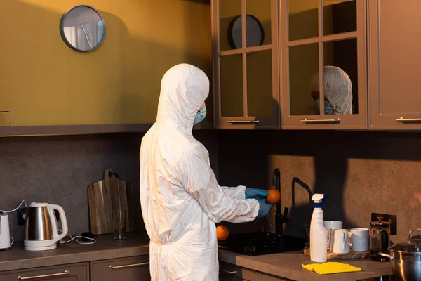 Вид сбоку человека в костюме хазмата, латексных перчатках и медицинской маске, моющей апельсин на кухне — стоковое фото