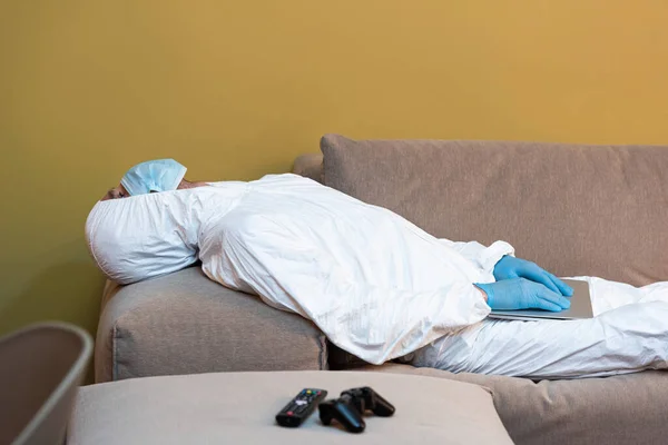 KYIV, UKRAINE - APRIL 24, 2020: Selective focus of man in hazmat suit and medical mask holding laptop near remote controller and joystick on couch — Stock Photo