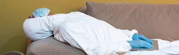 Panoramic shot of man in medical mask and hazmat suit holding laptop on couch — Stock Photo