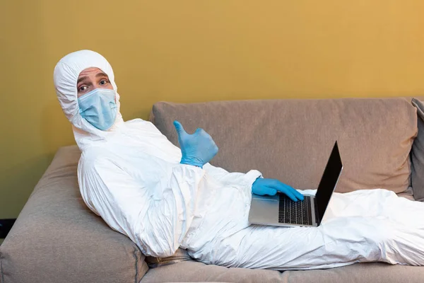 Vista lateral del hombre con máscara médica y traje de materiales peligrosos que muestra como signo en la cámara mientras sostiene el ordenador portátil en el sofá - foto de stock