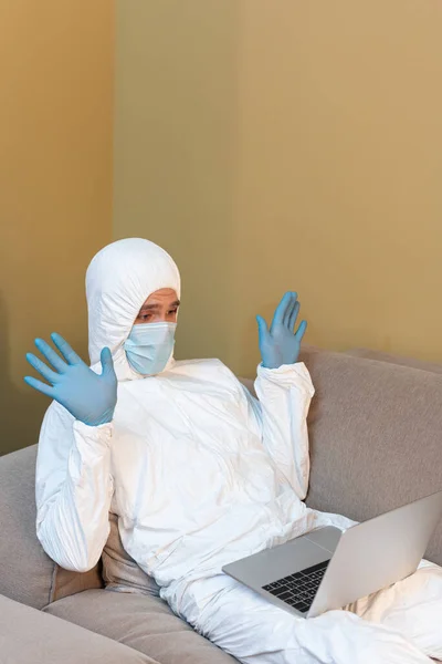 Man in hazmat suit and medical mask waving hands during video call on laptop — Stock Photo