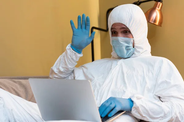 Selective focus of man in latex gloves, medical mask and hazmat suit having video call on laptop at home — Stock Photo