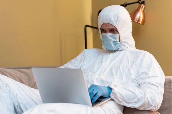 Selektiver Fokus des Mannes in medizinischer Maske und Schutzanzug mit Laptop auf der Couch — Stockfoto