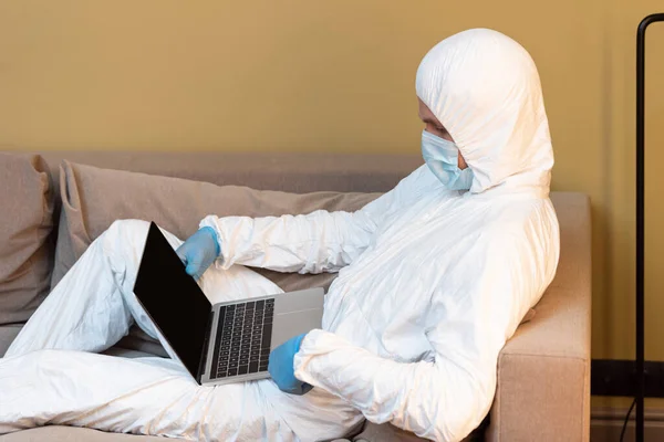 Вид збоку чоловіка в медичній масці, латексні рукавички і костюм з туманом, використовуючи ноутбук на дивані у вітальні — стокове фото